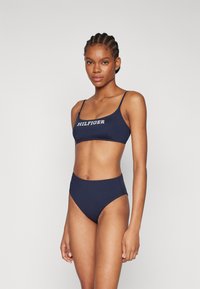 Tamsiai mėlynas bikini komplektas, turintis tankini tipo viršutinę dalį su plonomis petnešėlėmis ir "HILFIGER" logotipu, derinamas su aukštu juosmeniu apatiniais. Lygus audinio tekstūra.