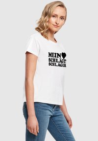 Balta īsām piedurknēm T-kreklā ar izteiksmīgu melnu tekstu "MEIN ♥ SCHLÄGT SCHLAGER." Klasisks apaļais kakls, viegli auduma kokvilna.