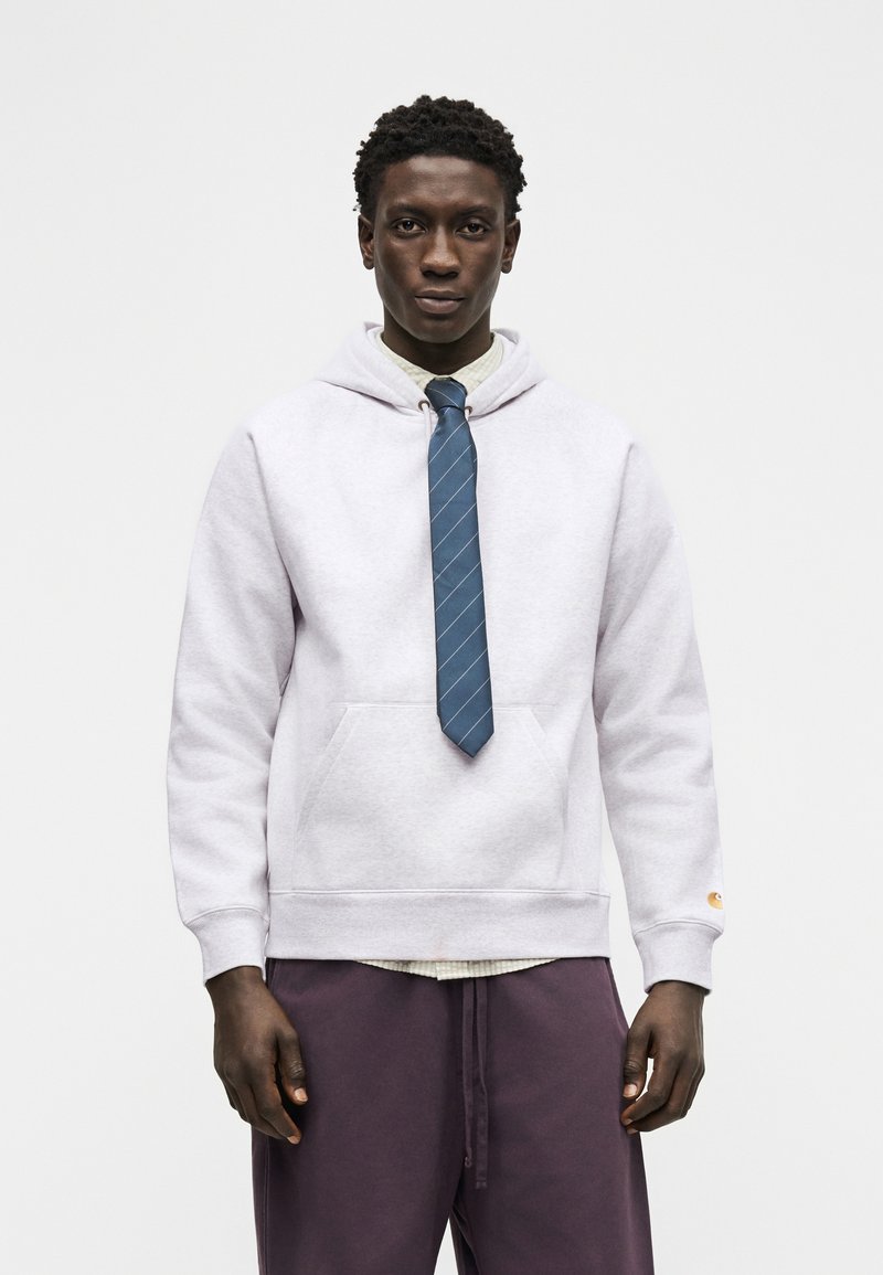 Man draagt een lichtgrijze hoodie over een overhemd met een blauw gestreepte stropdas, gecombineerd met donkerpaarse broek, staand tegen een witte achtergrond.