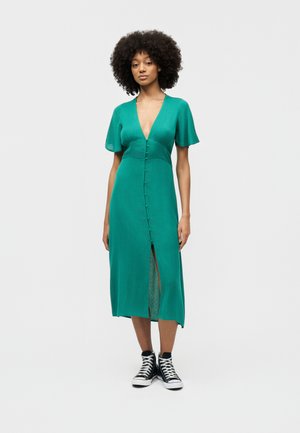 Vrouw met krullend haar, gekleed in een tealblauwe jurk met korte mouwen en knopen, en zwarte hoge sneakers, staand tegen een effen achtergrond.