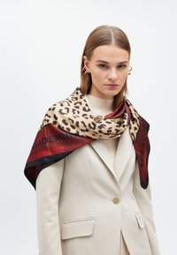 Seidenschal mit Leopardenmuster auf beigem Hintergrund, ergänzt durch rote und schwarze Akzente sowie geometrische Muster entlang der Kanten.