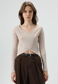 Lichtbeige geribbelde top met lange mouwen, een V-hals en wikkelontwerp, gecombineerd met bruine geplooide broek en een zwarte riem met een zilveren gesp.