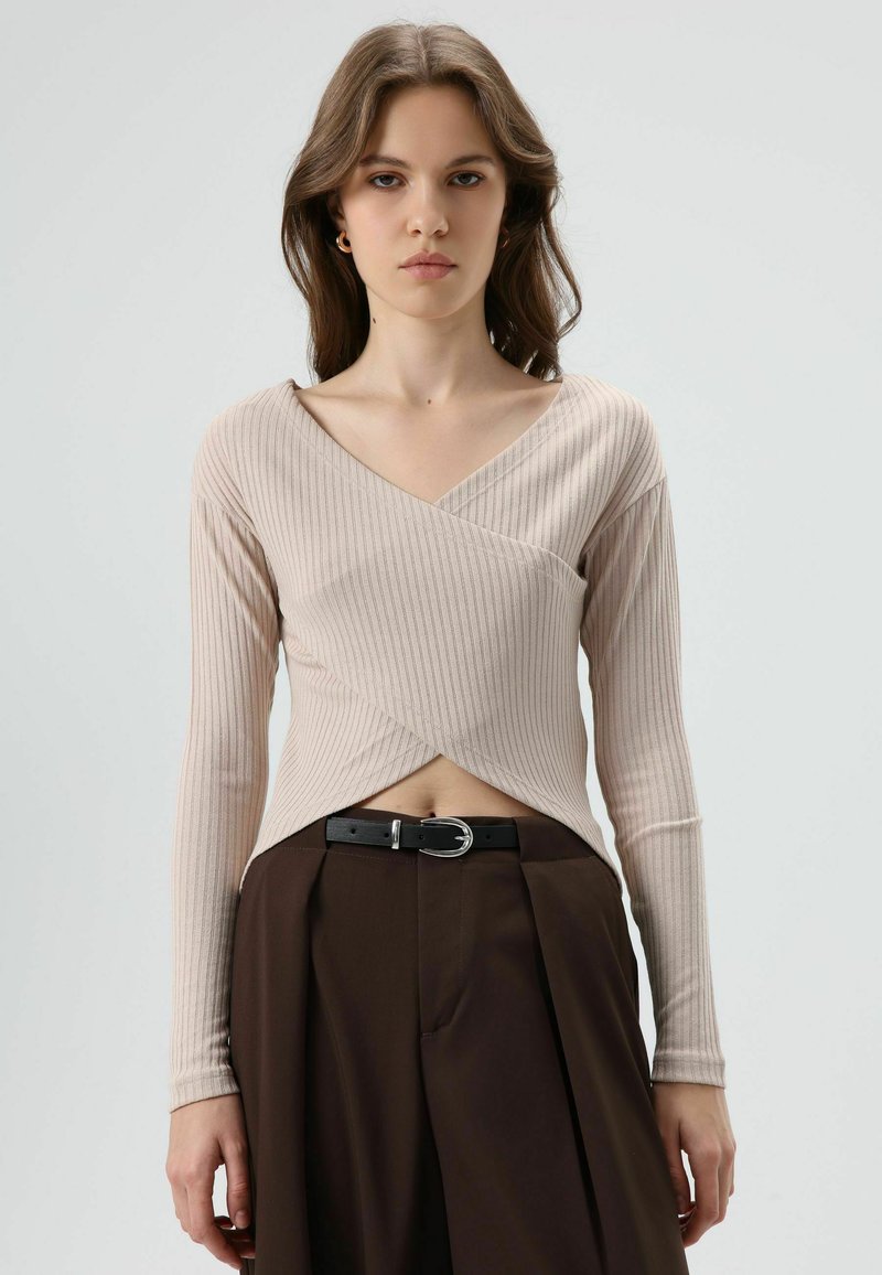 Lichtbeige geribbelde top met lange mouwen, een V-hals en wikkelontwerp, gecombineerd met bruine geplooide broek en een zwarte riem met een zilveren gesp.