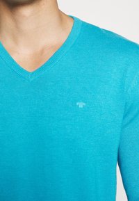 Pull turquoise à col en V en maille fine, avec un logo de marque discret sur le côté gauche. Texture lisse avec col côtelé.