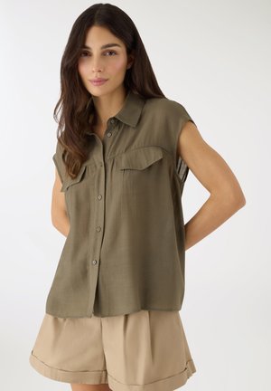 Donna con capelli lunghi e scuri che indossa una camicia oliva senza maniche con bottoni e pantaloncini beige a pieghe, in piedi con le mani dietro la schiena.