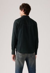 Camisa de mezclilla verde oscura con cuello abotonado, mangas largas y detalles de costura en el canesú; combinada con jeans azul desgastado.