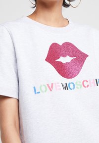 En person som bär en ljusgrå t-shirt med en stor glittrig grafik av rosa läppar och färgglad text "LOVE MOSCHINO" nedanför.
