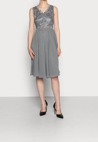 Robe grise avec un corsage en dentelle à motifs floraux, un décolleté en V, et une jupe fluide en chiffon. Portée avec des escarpins noirs à talons aiguilles.