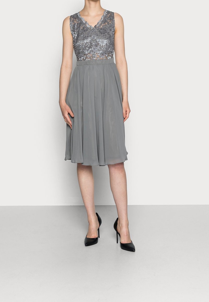 Robe grise avec un corsage en dentelle à motifs floraux, un décolleté en V, et une jupe fluide en chiffon. Portée avec des escarpins noirs à talons aiguilles.