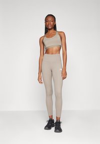 Soutien-gorge de sport beige et legging taille haute en matière lisse et ajustée. Des chaussures de sport noires avec des accents verts complètent la tenue.