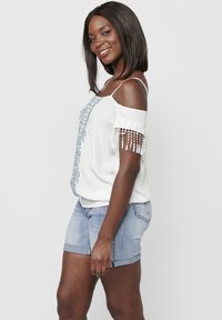 Top bianco, senza spalline, con ricami blu e dettagli di perline sulle maniche, abbinato a pantaloncini di jeans chiari. Tessuto liscio in tutto.