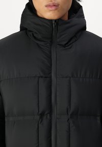 Schwarze Daunenjacke mit hohem Kragen, versehen mit einem gesteppten Muster und einem Reißverschluss vorne. Das Material wirkt glatt und leicht.