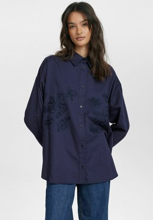 Navyblaues Hemd mit Knopfleiste und schwarzer floraler Stickerei auf der Vorderseite, aus leichtem Stoff gefertigt, mit Kragen und lockerer Passform.
