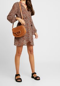 Robe fleurie marron à manches longues, taille cintrée et ourlet asymétrique. Sac bandoulière en cuir marron et sandales noires complètent la tenue.