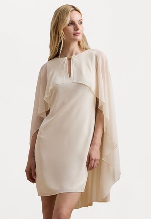 Beige mekko, jossa on kerroksellinen, puoliksi läpinäkyvä viitta ja avonainen kaula-aukko. Sujuva tekstuuri, polveen ulottuva, lempeillä laskostus yksityiskohdilla.