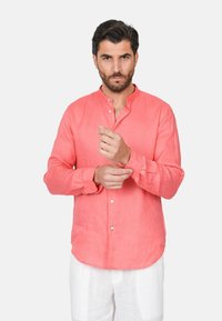 Camicia di lino corallo con colletto alla mandarina, maniche lunghe e polsini abbottonati. Texture liscia e vestibilità ampia, abbinata a pantaloni di colore chiaro.