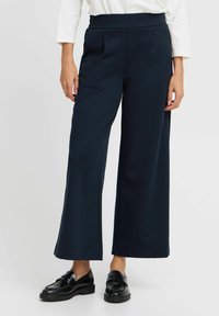 Pantaloni larghi blu navy con elastico in vita, caratterizzati da pieghe frontali e una superficie liscia, abbinati a mocassini neri.