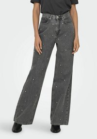 Pantalones vaqueros de mezclilla negra con piernas anchas, que presentan una cintura alta y pequeños adornos plateados esparcidos por toda la prenda. Bolsillos traseros lisos, diseño clásico.