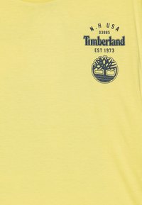Dzeltena kokvilnas T-krekls ar tumši zilu Timberland logotipu un koka simbolu, kā arī tekstu "N.H. USA 03885 EST 1973". Gluda tekstūra, īsas piedurknes.