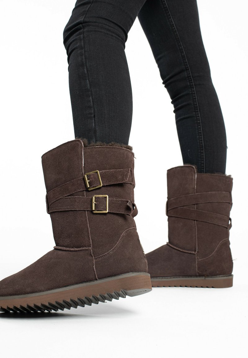 Botas de tobillo de ante marrón con dos correas cruzadas y hebillas. Suela de goma texturizada con un patrón dentado. Forro de forro polar visible en la parte superior.