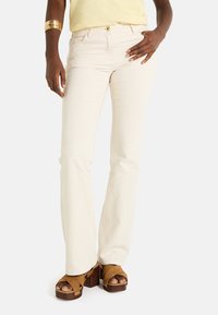Jeans de denim de corte slim en un beige claro con una textura sutil, cinco bolsillos y un cierre de botón único, combinados con sandalias de plataforma marrones.