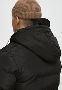 Doudoune noire avec capuche, présentant une texture brillante et des coutures visibles. Bonnet en tricot marron avec un motif côtes.