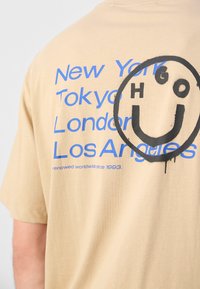 T-shirt beige en coton avec texte bleu listant des villes et grand graphique de smiley noir au dos. Manches courtes, coupe décontractée.
