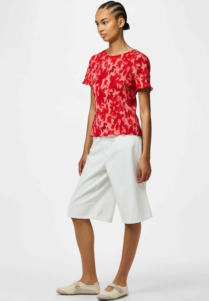 Femme debout portant un haut rouge à manches courtes fleuri, un short large blanc à hauteur des genoux, des chaussures Mary Jane beiges et les cheveux tressés coiffés en arrière.