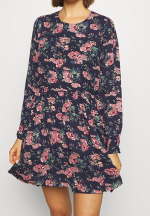 Femme portant une robe bleu marine à manches longues avec un motif floral rose et vert, tenant légèrement l'ourlet.