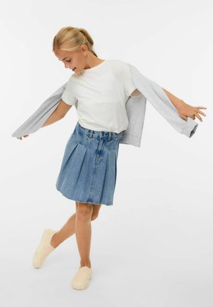 Jupe en denim bleu clair avec des plis, associée à un t-shirt blanc et un cardigan gris, complétée par des chaussures slip-on beiges.