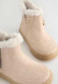 Beige suede ankelstøvler med en fluffy hvid foring, syet detaljer og strukturerede gummisåler. Lynlås på siden for nem påtagning.