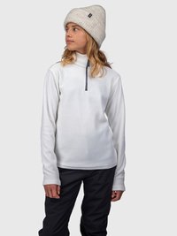 Una giovane persona che indossa un fleece bianco con zip, un berretto a maglia beige e pantaloni neri, che guarda di lato contro uno sfondo semplice.