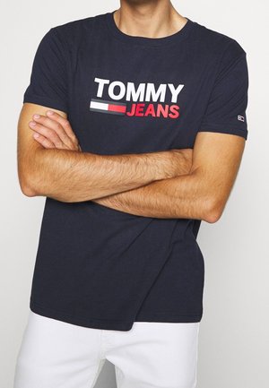 Mężczyzna w granatowej koszulce Tommy Jeans z logo i białych spodniach, z rękami skrzyżowanymi na piersi, prezentujący tors i górną część nóg.