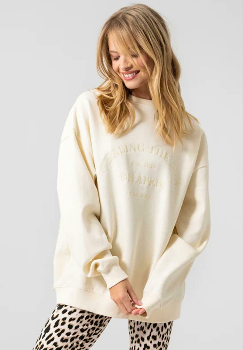 Sudadera oversized crema con cuello redondo y puños de canalé, que presenta texto en relieve. Combinada con leggings de estampado de leopardo.