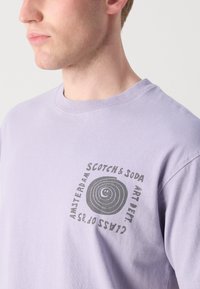 Lilla bomull t-skjorte med en sirkulær grafisk trykk i mørk grå, med "Scotch & Soda" og "Amsterdam" tekst vist tydelig.