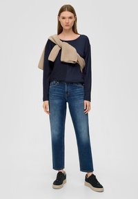 Navy-blauer Langarmoberteil, beiger Pullover lässig über die Schultern gelegt, kombiniert mit mittelhohen blauen Jeans und schwarzen Sneakers mit kräftigen Sohlen.