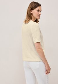 Krátký rukáv krémového topu s texturovanou tkaninou, kulatým výstřihem a ohrnutými rukávy, kombinovaný s bílými kalhotami. Zpětný pohled ukazuje jemný design.