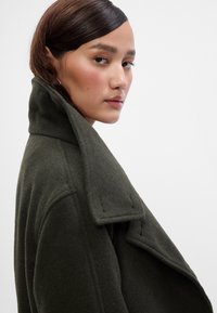 Casaco de lã verde oliva com um colarinho largo e elevado, ombros estruturados e linhas limpas. Textura suave com costuras visíveis.