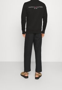 Felpa nera con scritta bianca "TOMMY HILFIGER" sul retro, abbinata a pantaloni neri larghi e sandali marroni. Materiale in cotone, design casual.