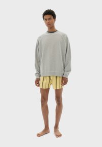 Grå översize sweatshirt med rund halsringning, kombinerad med gula shorts med ränder. Modellen står barfota mot en enkel vit bakgrund.