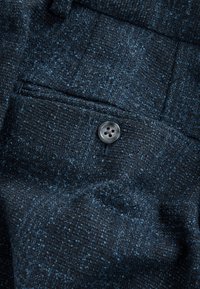 Tela tejida de color azul oscuro con una superficie texturizada. Un solo botón gris detalla y realza el diseño. Cuenta con un pequeño bolsillo.