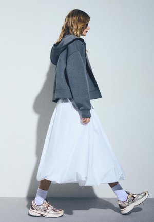 Donna che cammina con felpa con cappuccio grigia, gonna bianca voluminosa, calzini bianchi e sneakers beige su sfondo chiaro uniforme.