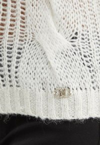 Maglione bianco lavorato a maglia con un'orditura larga e aperta, orlo a coste solido e un accento con logo argentato vicino al fondo.