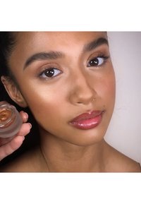 Clear cosmetic jar held next to a model's face. Model has a tan complexion, glossy lips, and defined eyebrows. Jar contains a bronze cream.