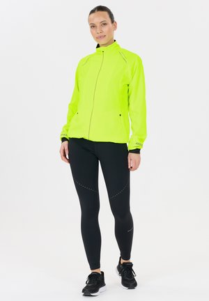 Fluorescerande gul jacka med dragkedja framtill och hög krage, kombinerad med svarta leggins med prickiga mönster längs sidorna och svarta sneakers.