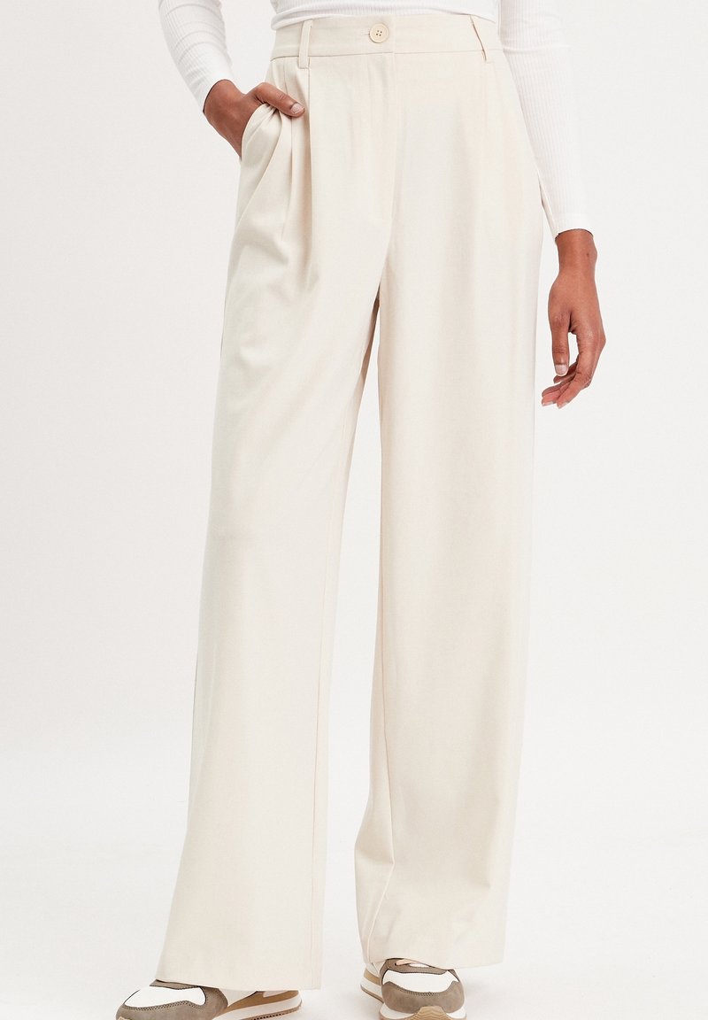 Pantalon à jambes larges en tissu doux couleur crème, avec des plis sur le devant et des poches latérales. Finition avec une fermeture à bouton à la taille.