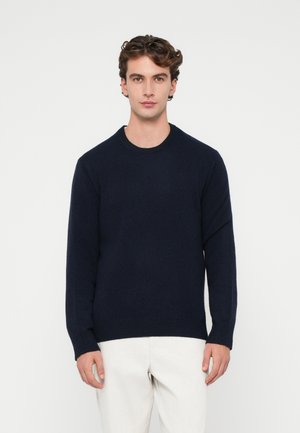 Pull col rond bleu marine à manches longues, avec des poignets et un ourlet côtelés, en tissu tricoté doux et texturé. Porté par-dessus un pantalon clair.
