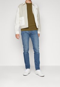 Lättviktsjacka med färgblockering i beige och vitt, med bröstficka, kombinerad med en olivgrön t-shirt och blå jeans. Vita sneakers.