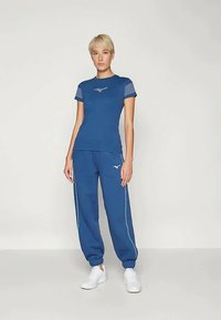 Ženski model stoji v modri atletski majici in hlačah za tek ter belih supergah na enobarvni podlagi.