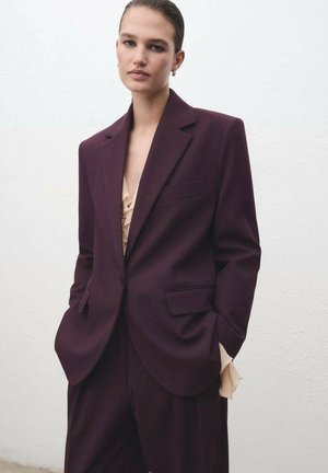Blazer entallado de color púrpura oscuro con solapas de muesca, un bolsillo y corte relajado, combinado con una blusa nude con volantes. Textura suave.